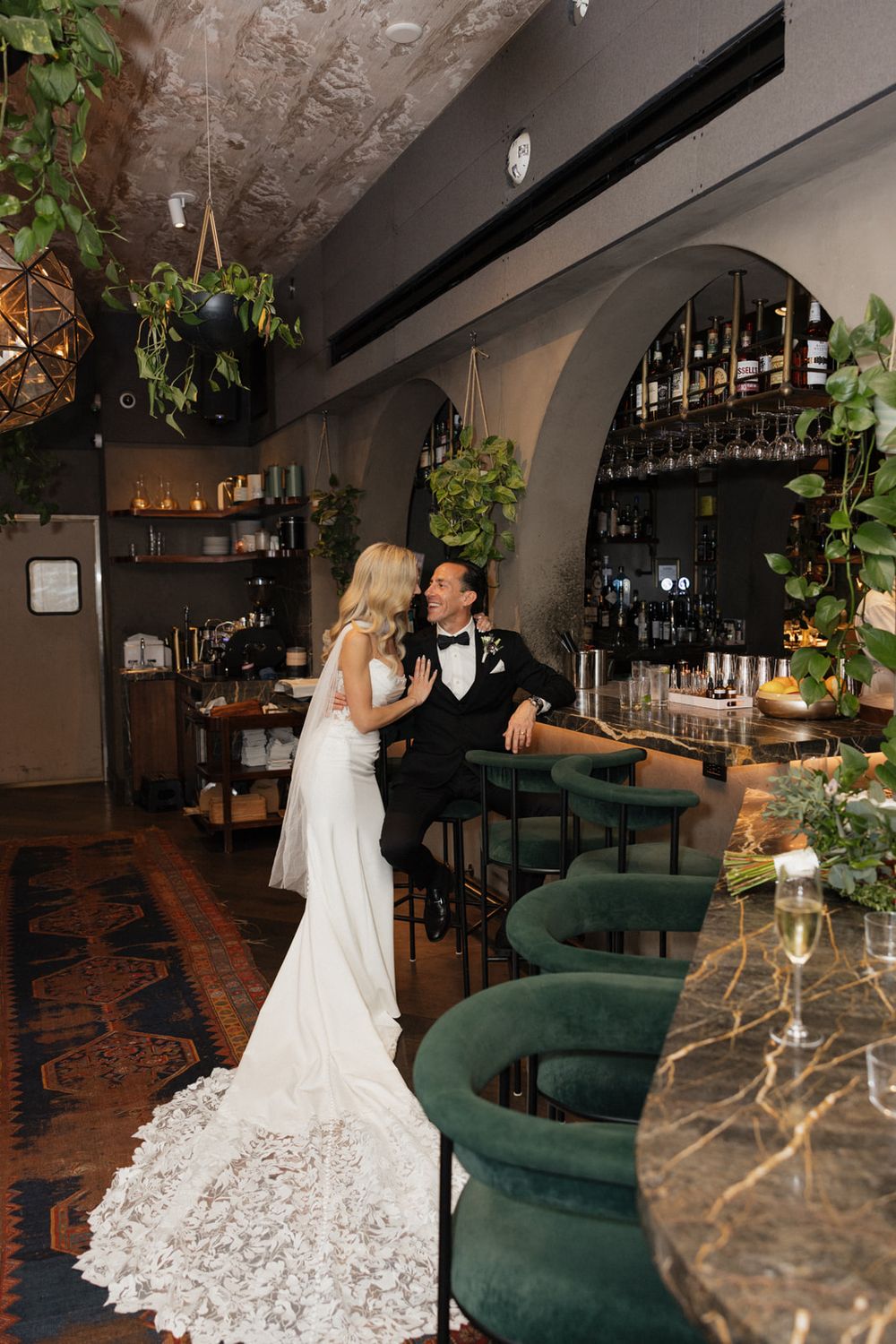 Elegant wedding venue with emerald velvet seating, dark wood bar, and hanging plants creating a romantic atmosphere.