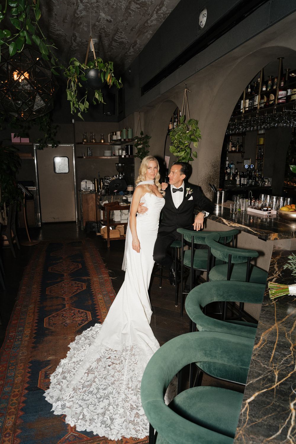 Newlyweds pose at an elegant bar with green velvet stools and a dramatic lace wedding dress train.