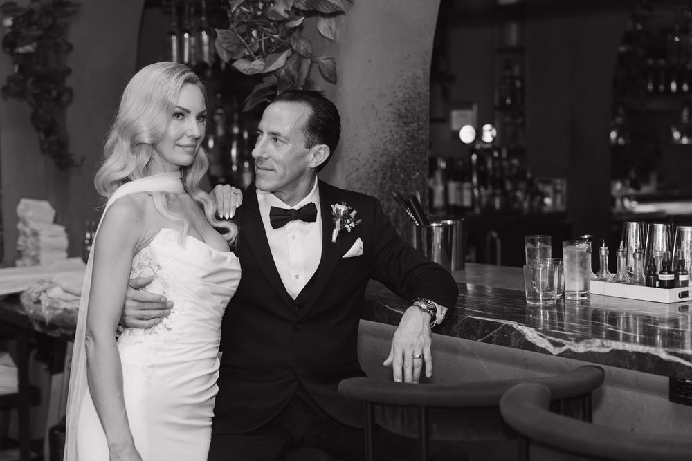 A glamorous black and white wedding photo shows a couple posing at a bar in formal attire.