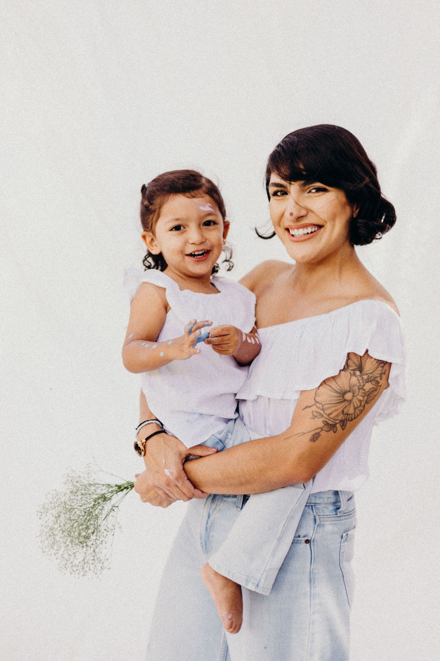 A woman in a white top and jeans holds a small child wearing a light colored outfit against a white background.