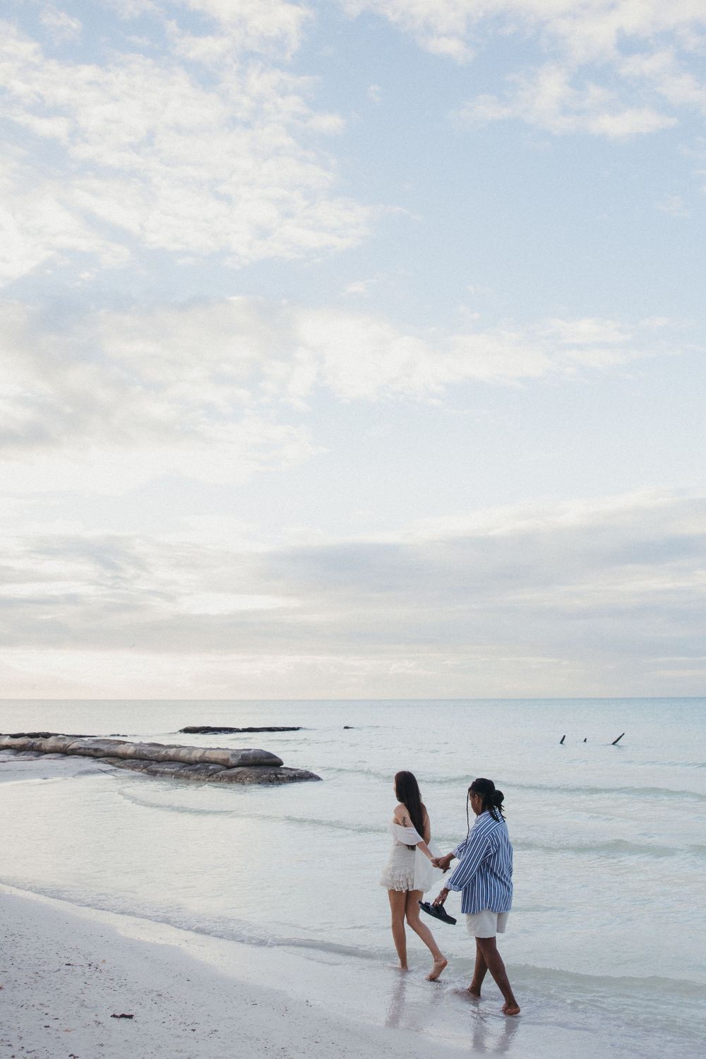 A gay couple walk along a serene beach in Holbox with white sand and turquoise waters and cloudy skies in the background.