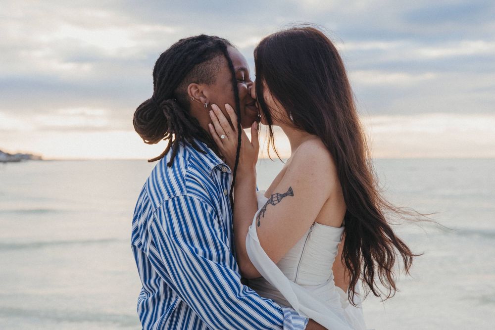 Couple embraces while laughing together because she said yes to their surprise proposal on a sandy beach with ocean waves in the background