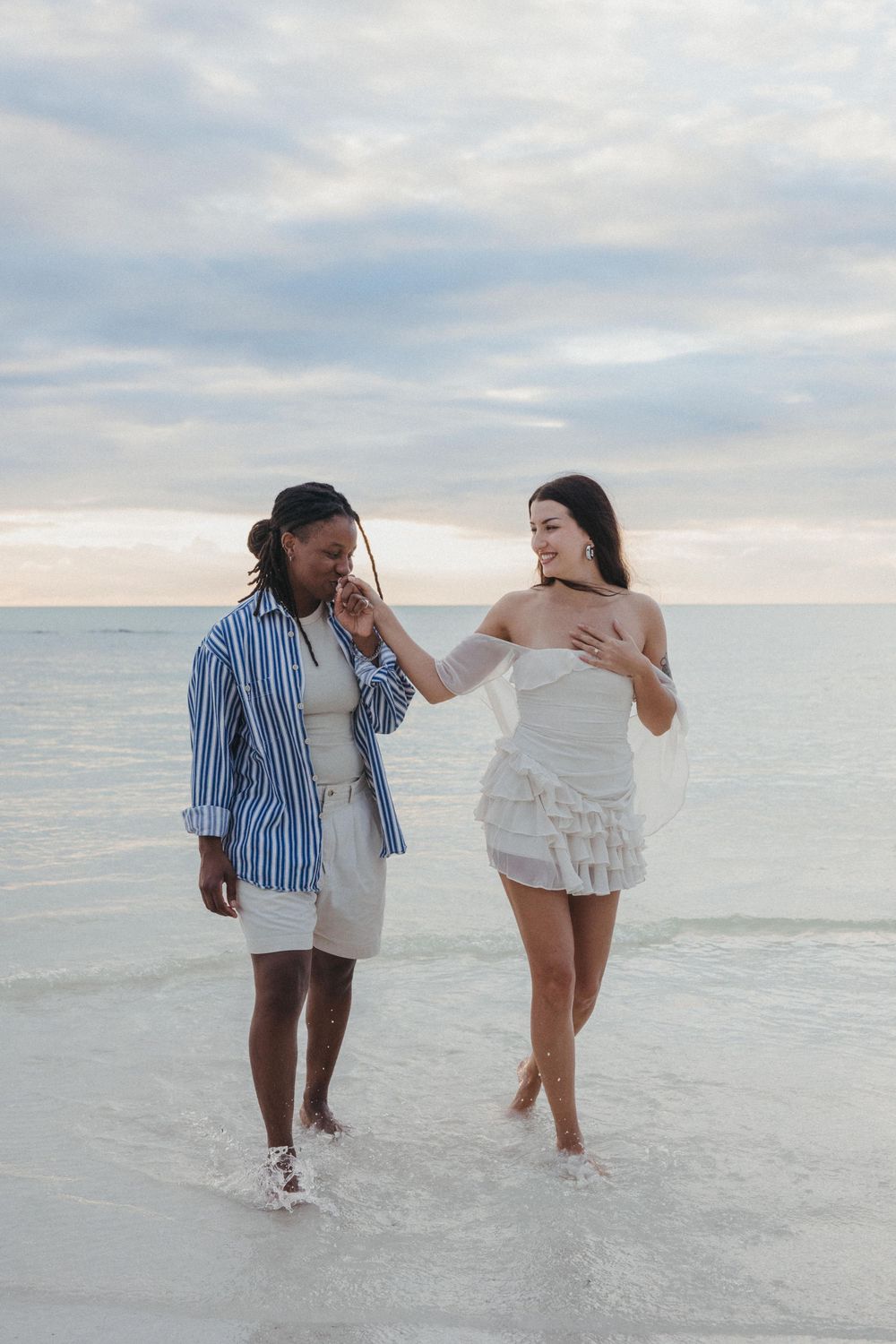 Two women walking along the beach in Holbox as one gently kisses the engagement ring after their surprise proposal at sunset.