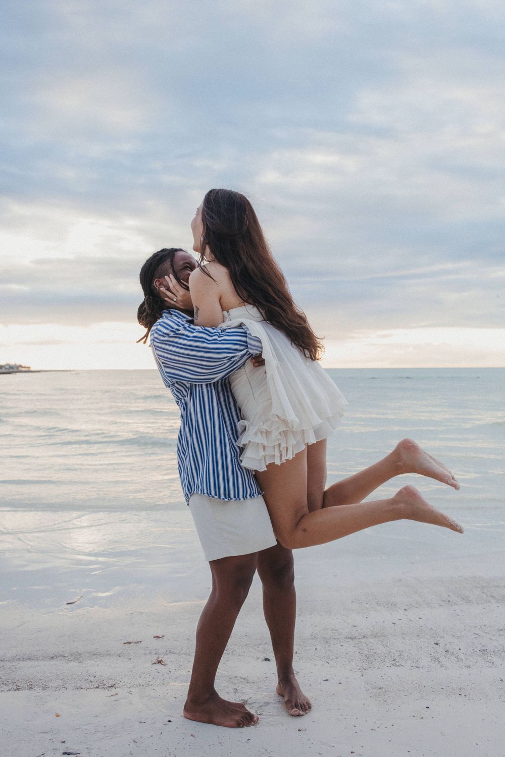A romantic couple embraces after proposing on a serene beach at Holbox sunset with white sand and calm ocean waters in the background.