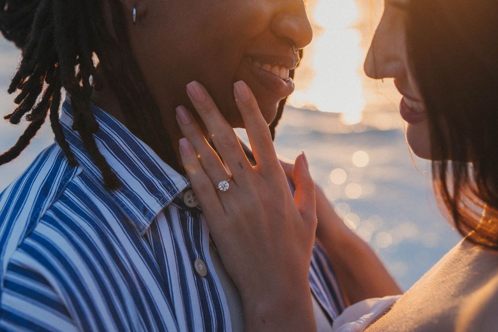 Close-up of a gay couple holding each other’s faces, foreheads nearly touching, with an engagement ring visible and the Holbox sunset reflec