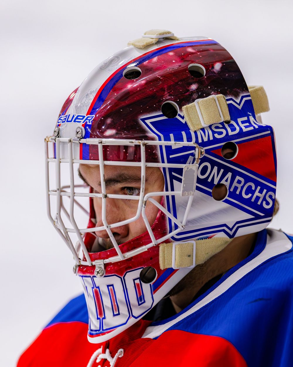 Close-up of a hockey goalie wearing a Lumsden Monarchs decorated mask during an ice hockey game.