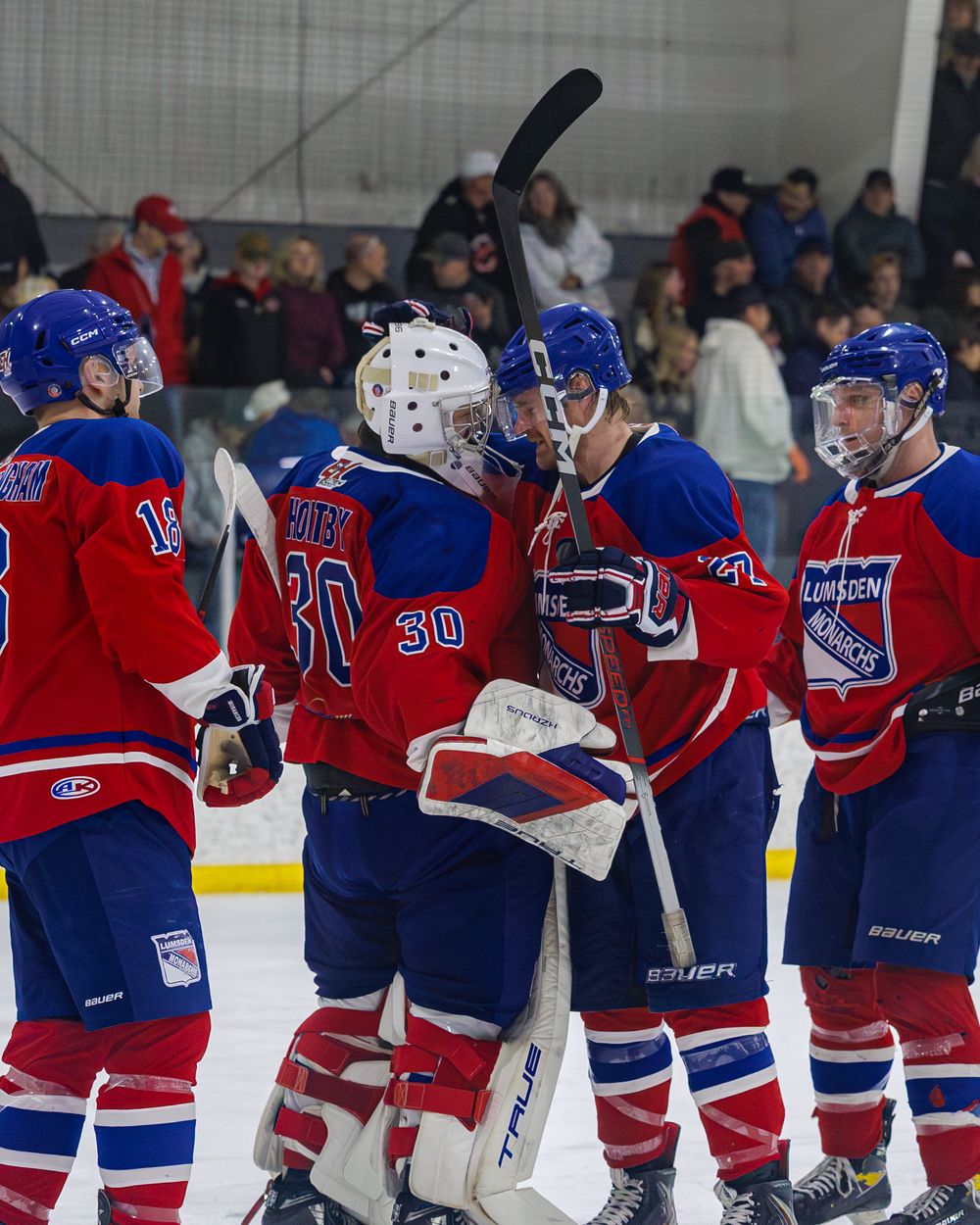 Red and blue hockey players celebrate on ice after a game, with goalie number 30 at center of the group.