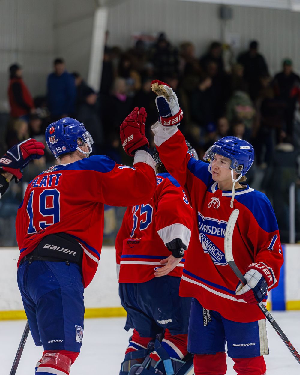 Youth hockey players in red and blue uniforms celebrate with high fives on the ice after a goal.