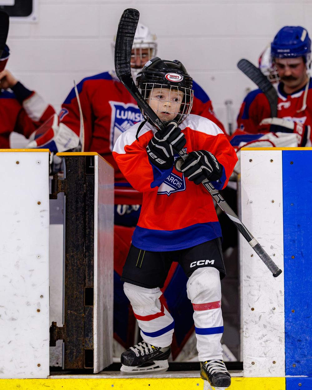 Young hockey player in Lumsden Monarchs jersey exits the bench with stick raised during a youth ice hockey game.