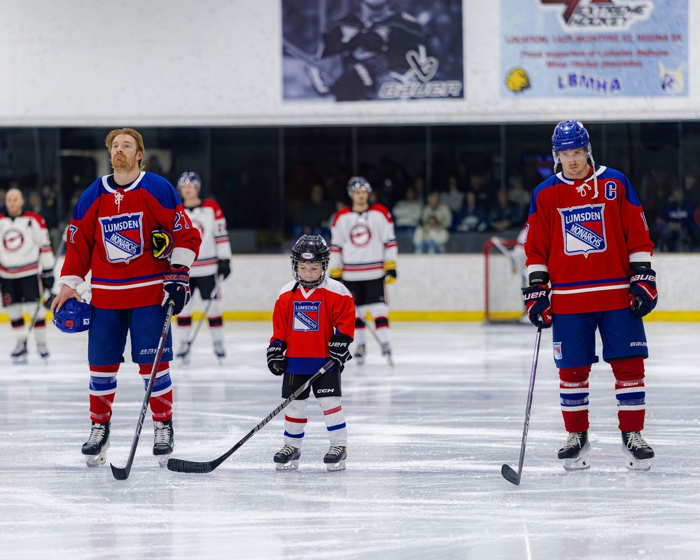 Two adult Lumsden Monarchs players stand with a young child hockey player during an ice rink ceremony.