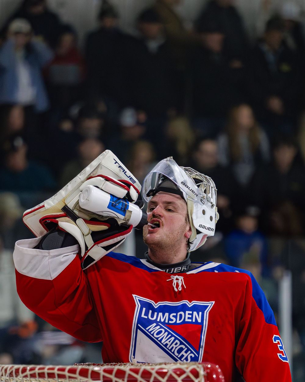 Lumsden Monarchs goalie removes helmet near the net while crowd watches during a competitive hockey game.