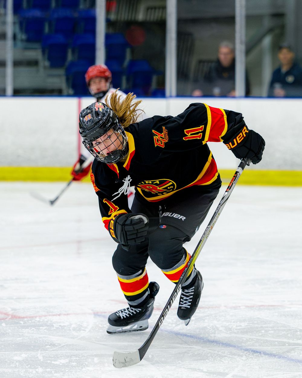 Female ice hockey player in black, red, and yellow Bauer uniform skating aggressively during a rink game.