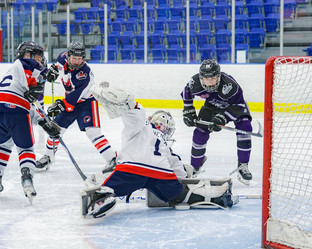Hockey goalie in red and white makes a save as players battle in front of the net during a game.