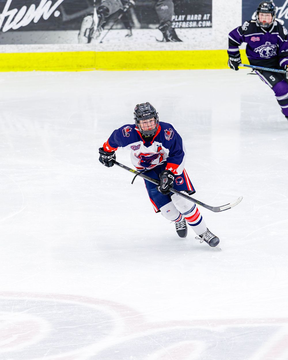 Young hockey player in red and blue uniform skates hard down the ice during a competitive game.