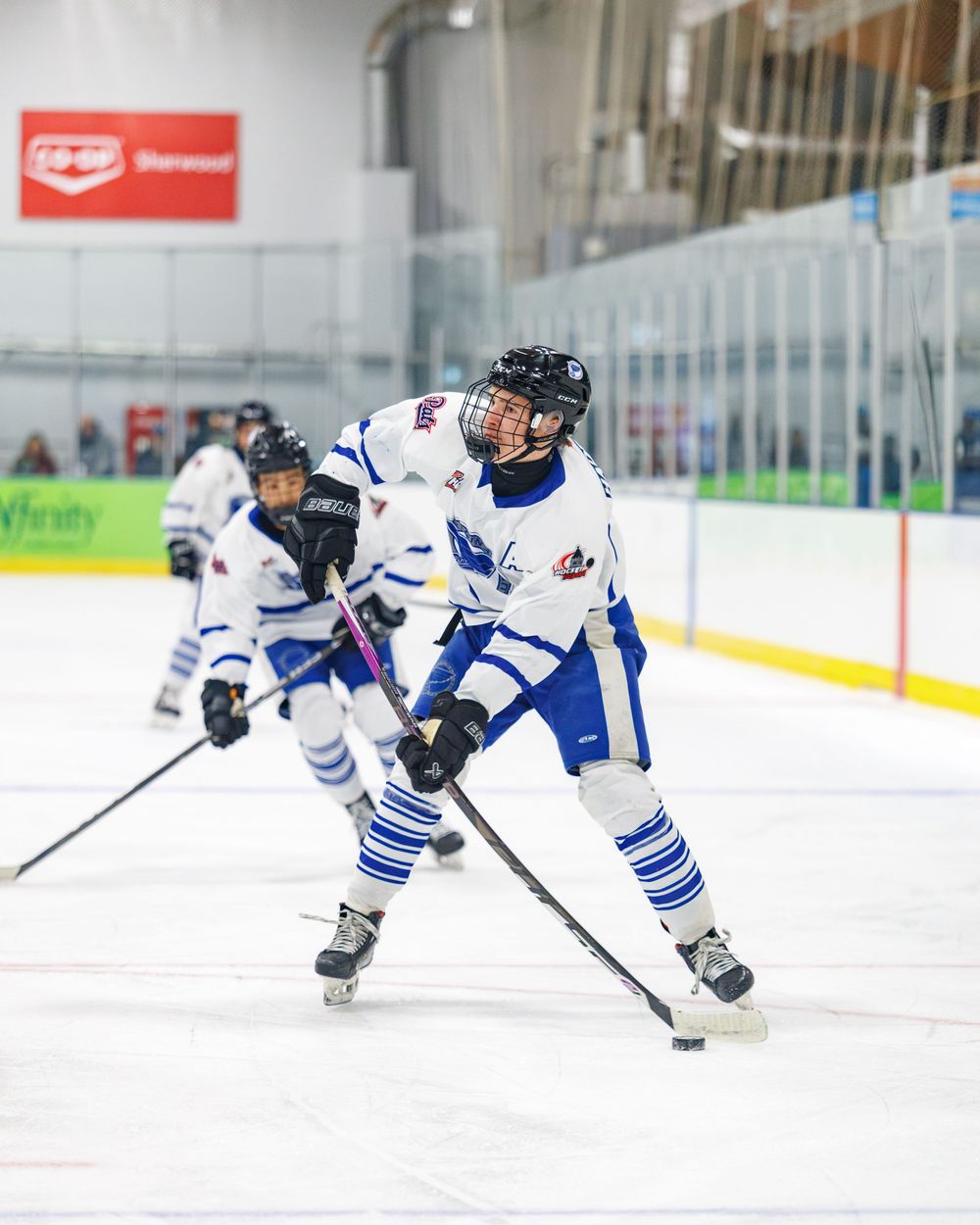 Youth ice hockey player in white and blue jersey handles puck during indoor rink game.