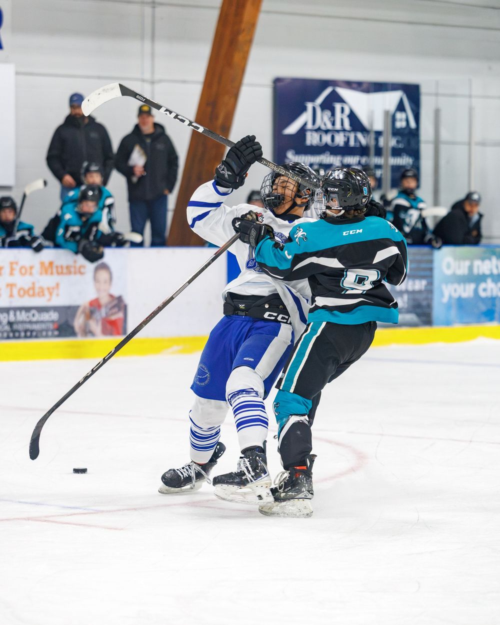 Two ice hockey players collide during a game, with spectators and a D&R roofing advertisement visible in the background.