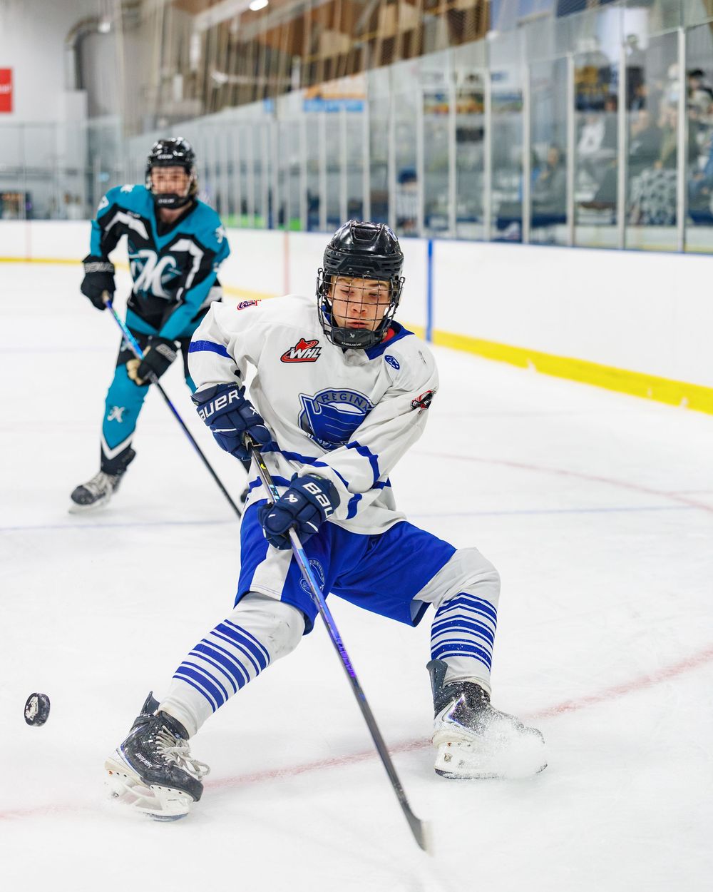 Young hockey player in white and blue uniform skating and shooting puck on indoor ice rink during game.