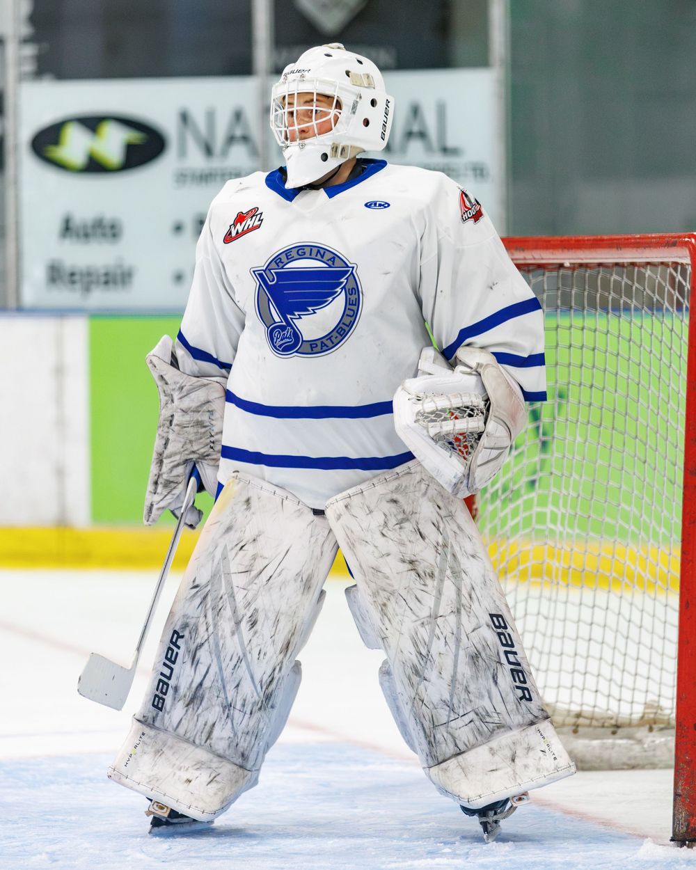 Ice hockey goalie in white and blue uniform stands ready in front of net on indoor rink.