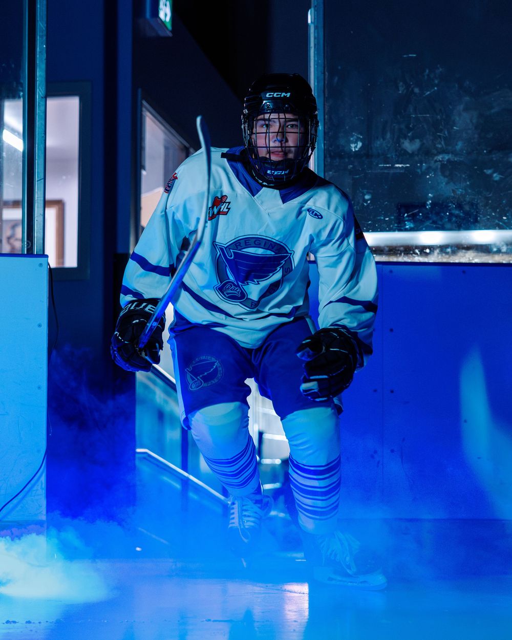 Young ice hockey player in white jersey emerges through blue smoke in dramatic rink entrance.