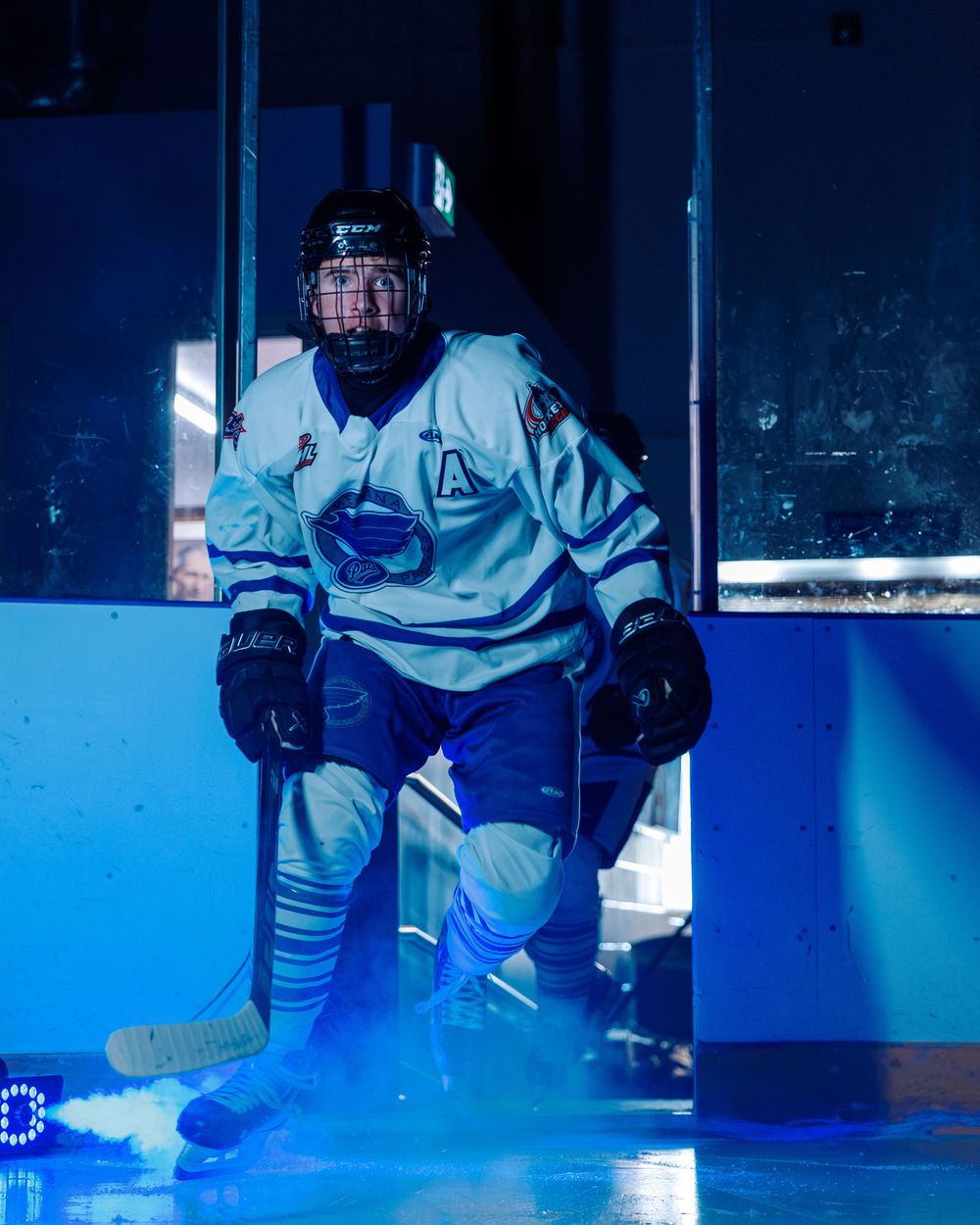 Hockey player in white jersey with letter A skates onto ice rink under dramatic blue lighting.