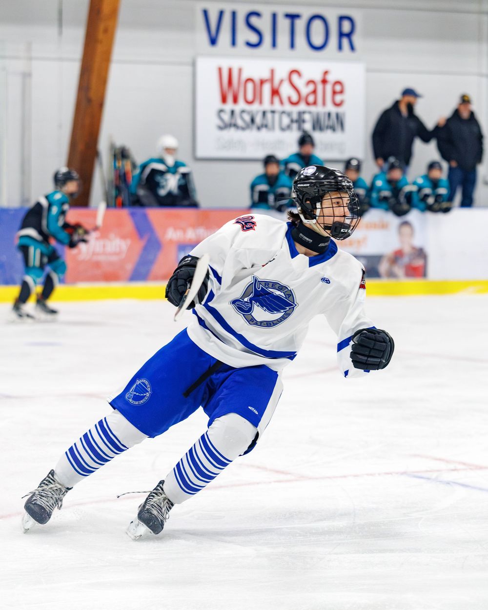 Youth hockey player in white and blue uniform skating on ice rink during a visitor game in Saskatchewan.