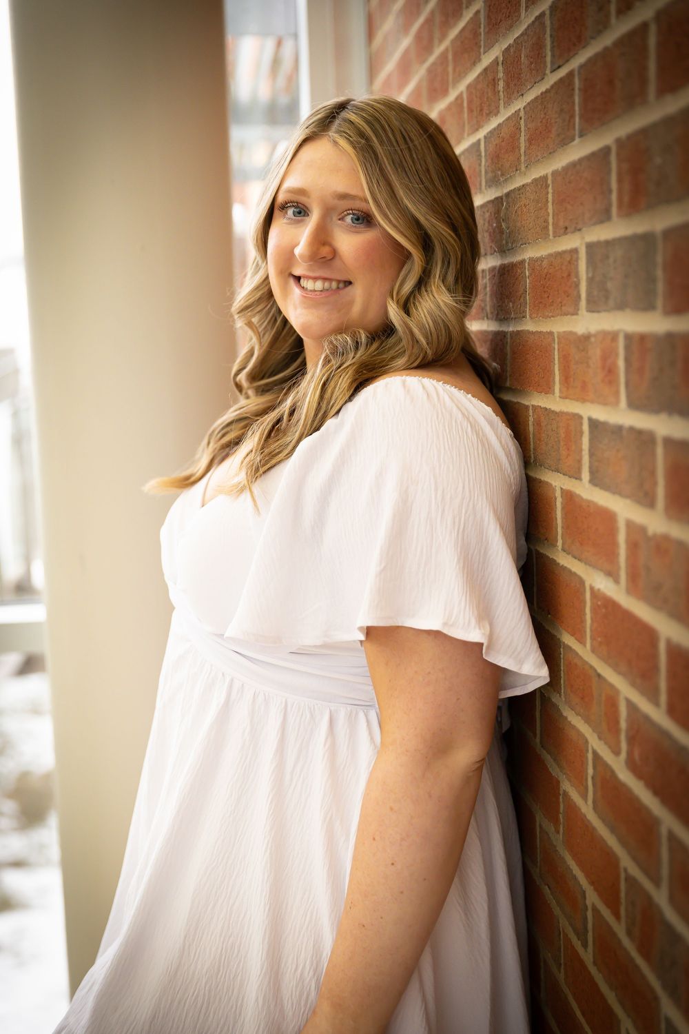 Young woman in white flutter-sleeve dress smiling while leaning against red brick wall in natural light.