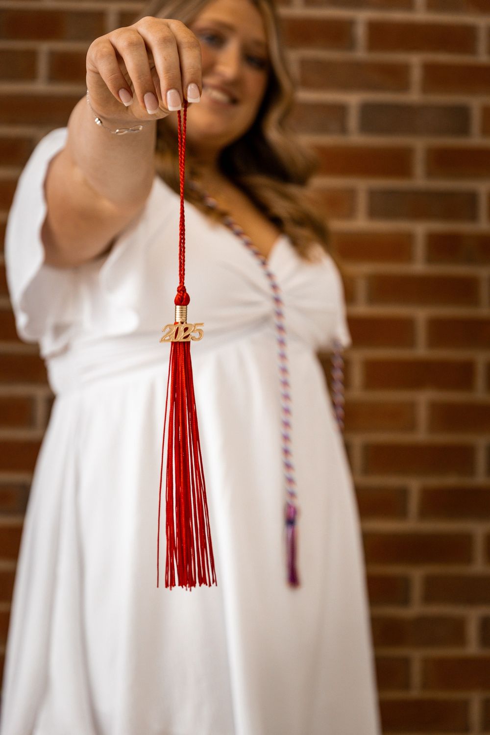 Graduate in white dress holding red 2025 graduation tassel toward camera with brick wall background.