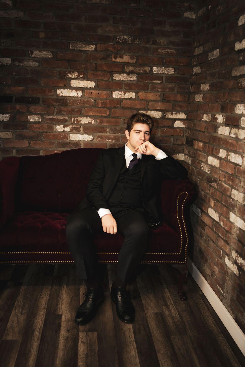 Young man in black suit sitting on red velvet sofa against brick wall in moody studio portrait.