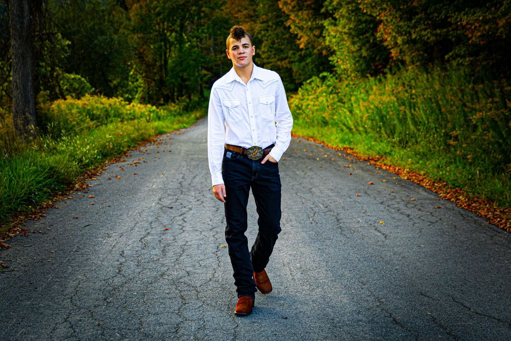 Young man in white shirt and dark jeans walks confidently down a rural tree-lined road at sunset.