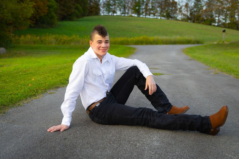 Smiling young man in white shirt and boots sits relaxed on a winding country road with green hills.