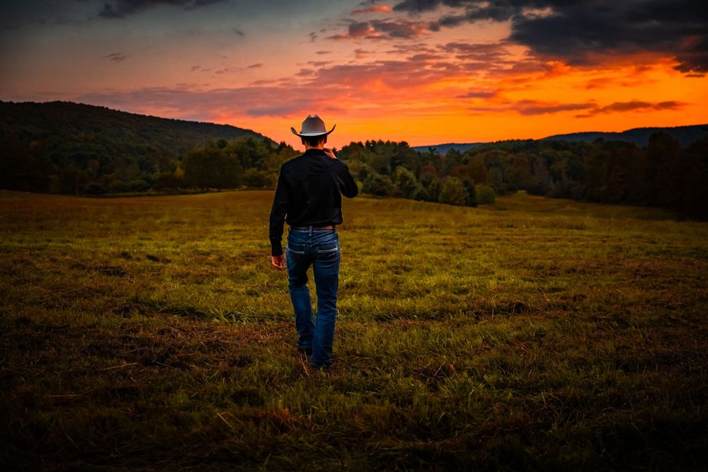 Cowboy in hat and jeans walks through green field toward vibrant orange sunset sky.