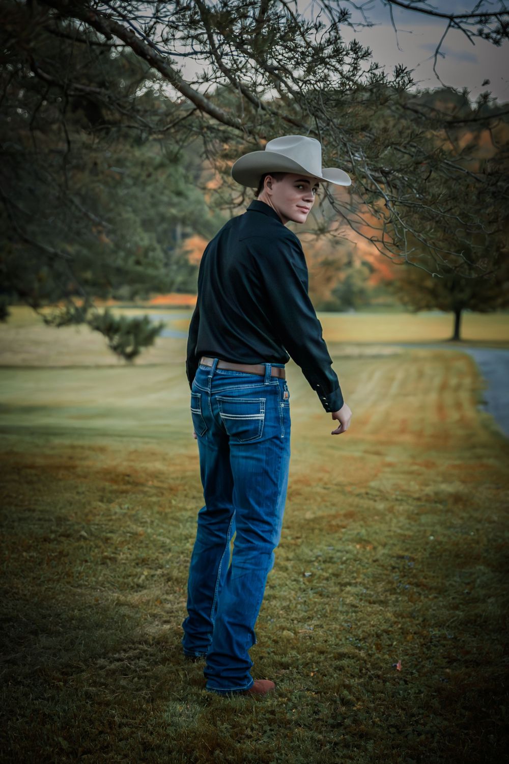 Young man in cowboy hat and black shirt poses outdoors at dusk near trees.