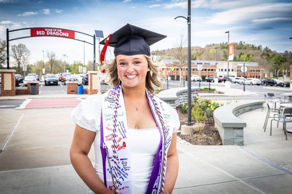 Cheerful female graduate wearing a decorated stole and cap smiles outside SUNY Red Dragons campus.