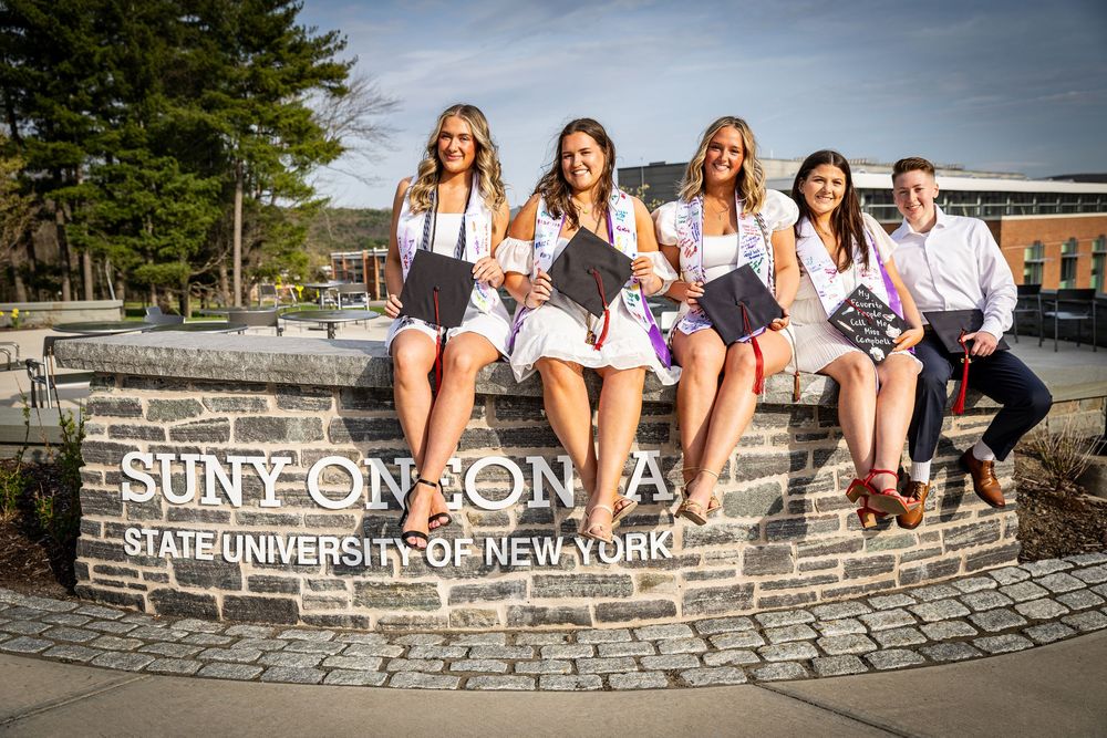 Five graduates sit on a SUNY Oneonta State University of New York stone sign holding diplomas.