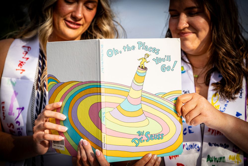 Two female graduates hold open a copy of Dr. Seuss Oh the Places Youll Go book at graduation.