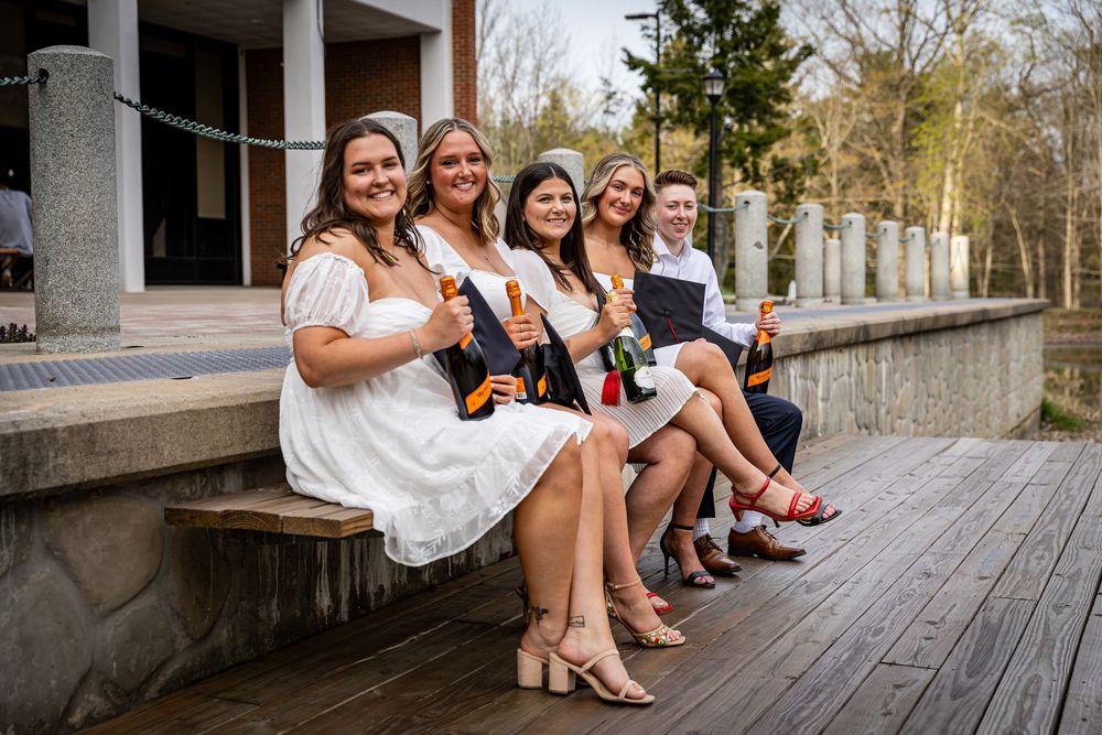 Five young women in white dresses sit on wooden steps smiling, holding champagne bottles at a graduation celebration.