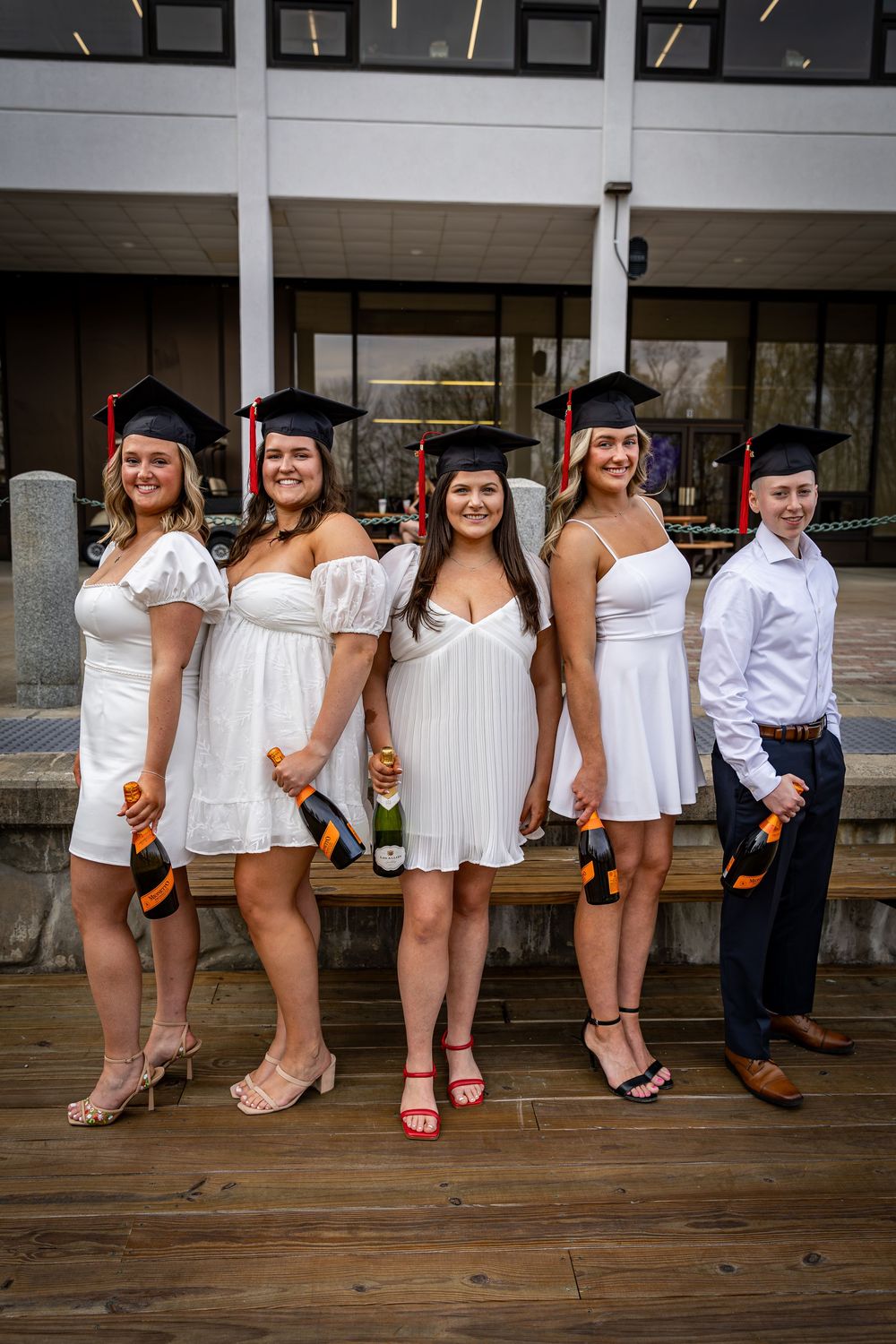 Five graduates in white dresses and black caps pose outdoors holding champagne bottles to celebrate graduation day.
