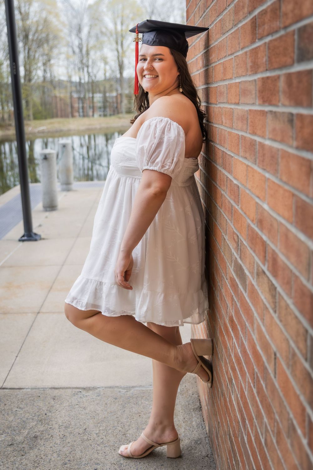 Smiling young woman in white off-shoulder dress and graduation cap leans against brick wall by a pond.