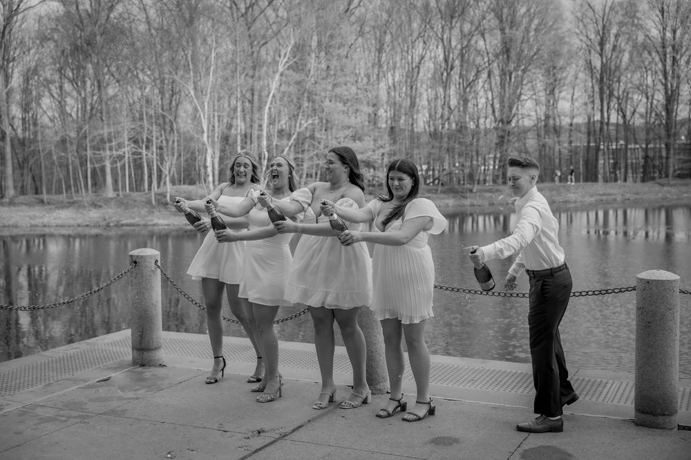 Black and white photo of five graduates in white dresses popping champagne bottles by a scenic pond.