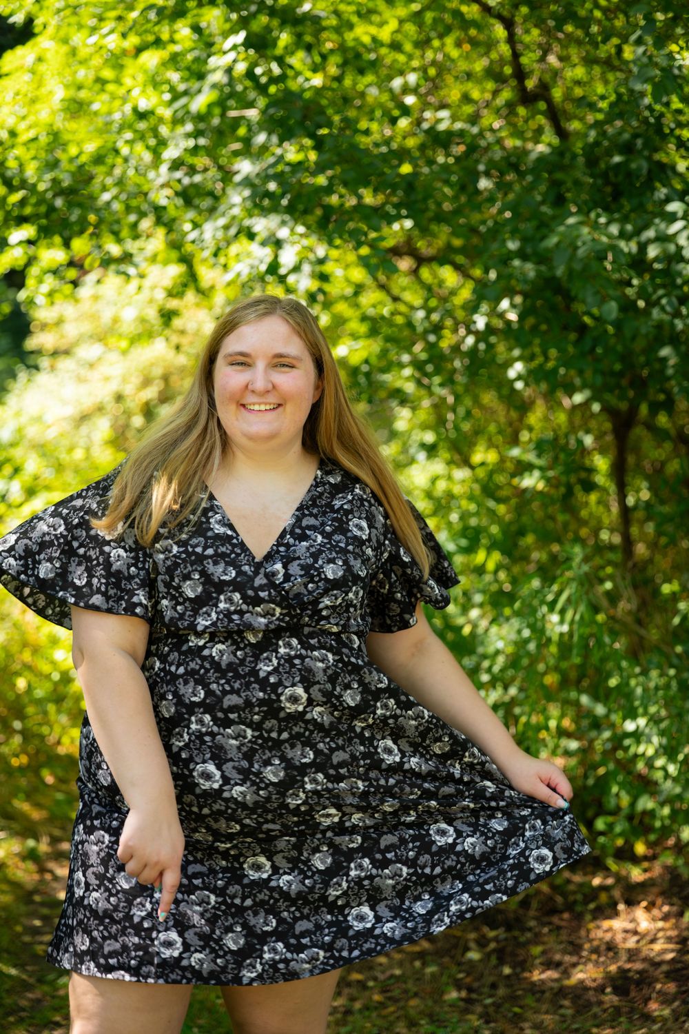 Cheerful blonde woman in floral dress smiles on sunlit forest path.