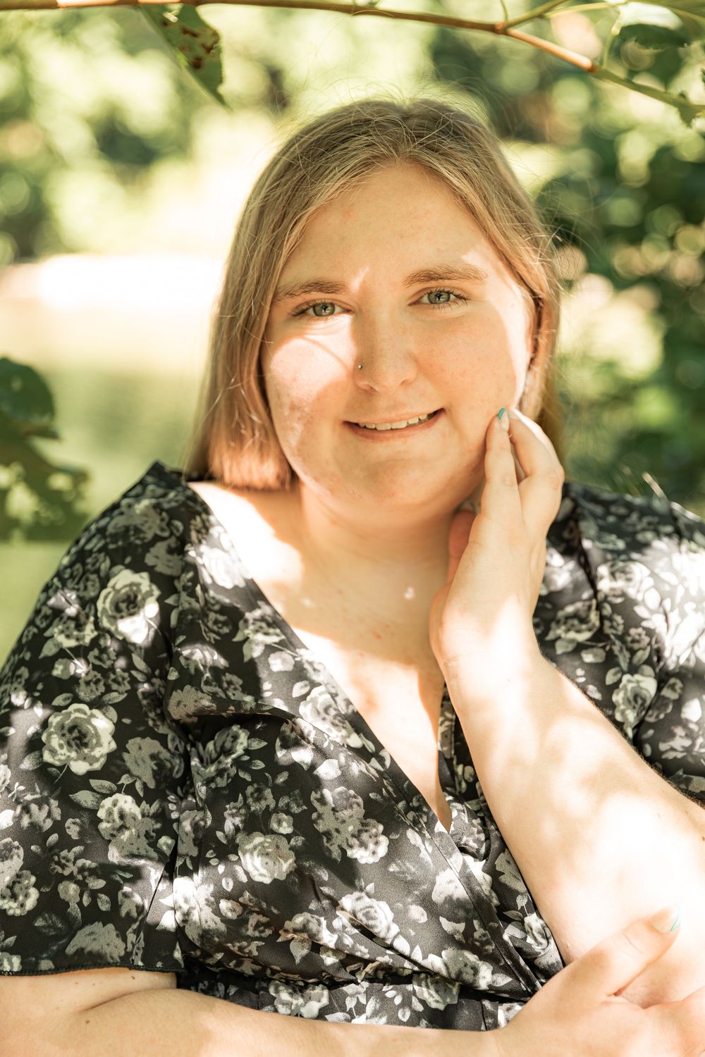 Blonde woman in floral dress smiles softly under dappled sunlight near green leaves.