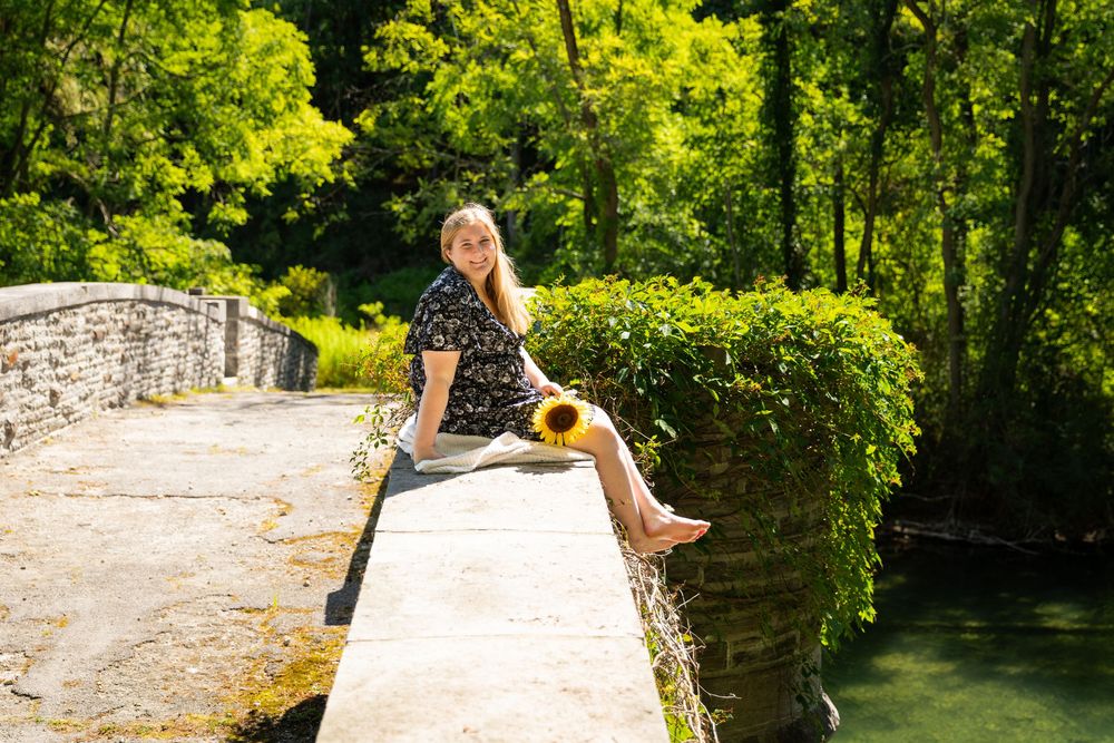 Young woman holding sunflower sits on ivy-covered stone bridge over calm green water.