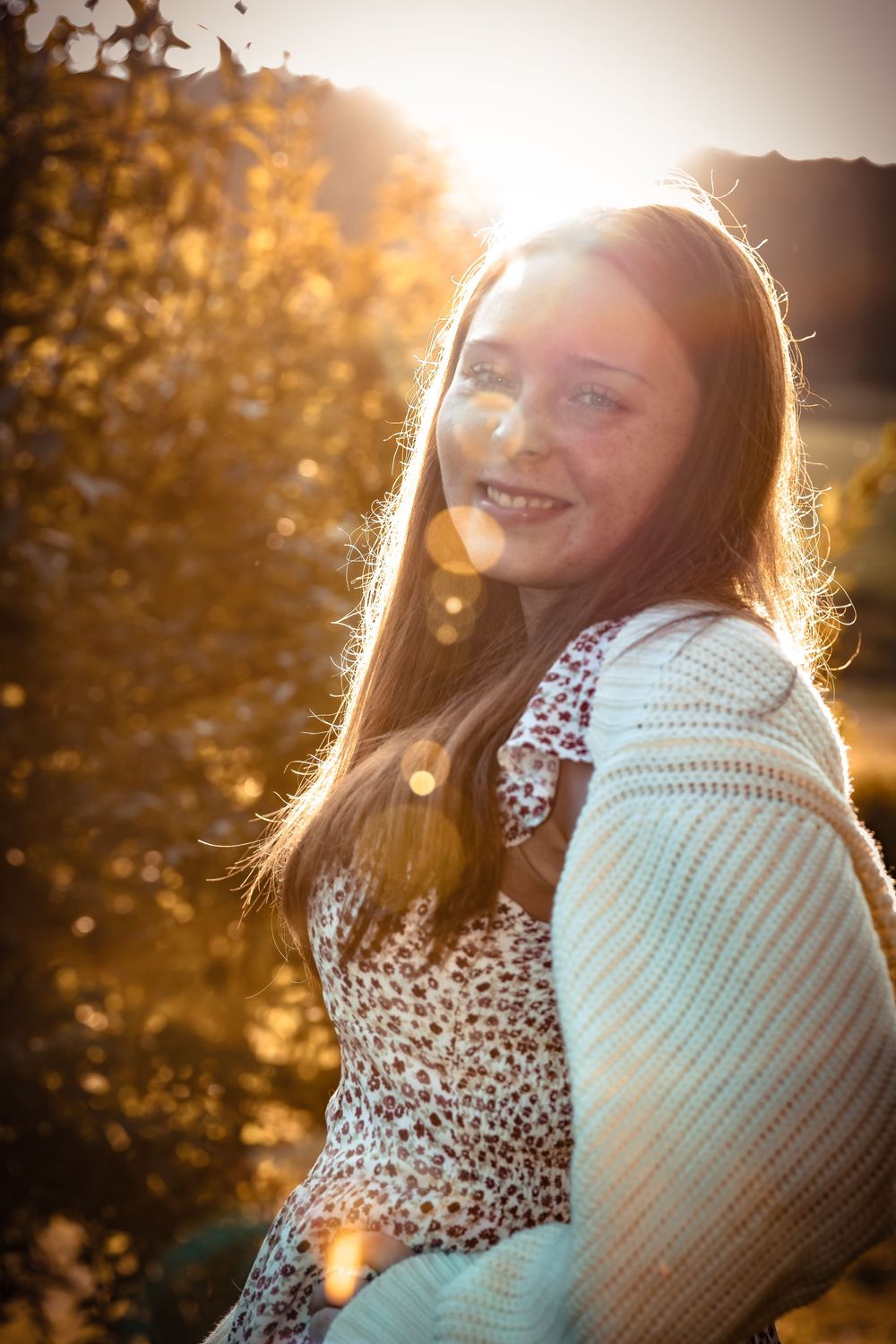 Teenage girl with long red hair and freckles smiling in golden sunset light wearing a floral dress and white cardigan.