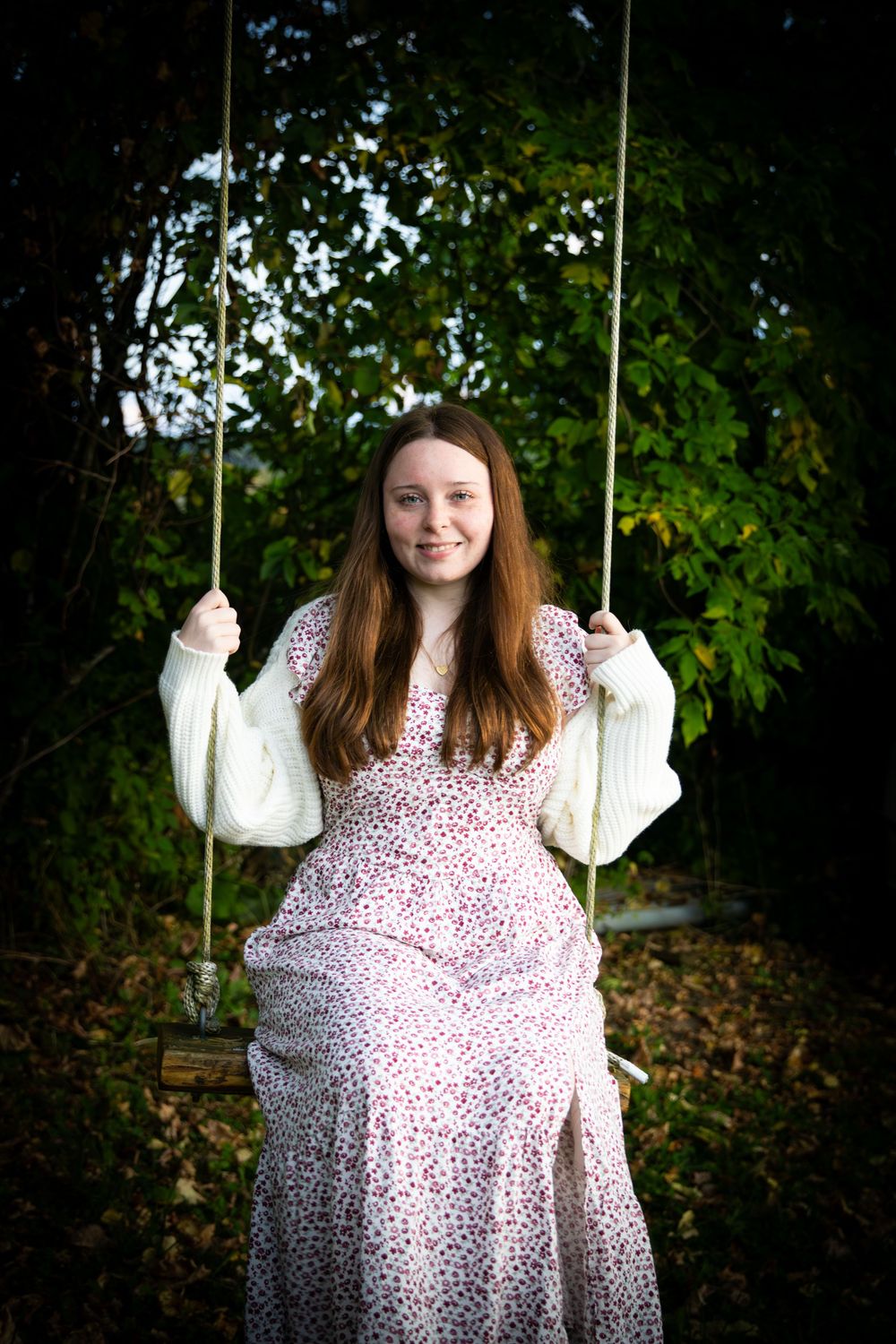 Smiling young woman with long auburn hair in a floral dress sitting on a rope swing surrounded by autumn trees.