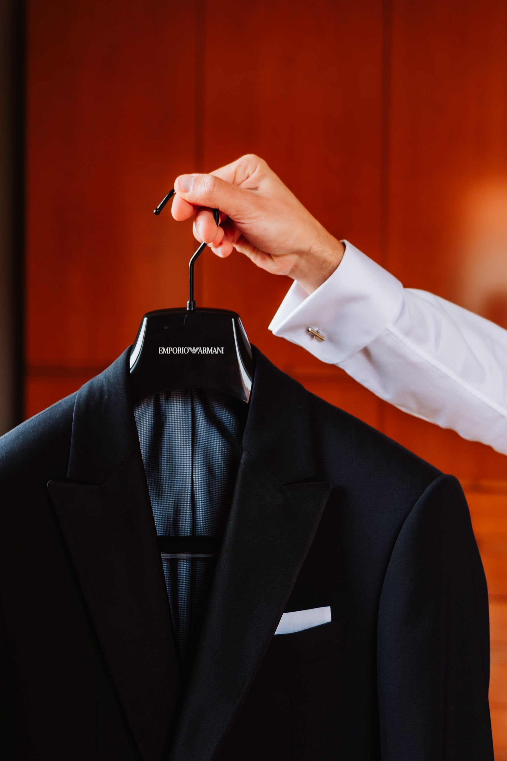 A hand places a black suit jacket on a wooden hanger against a warm red-toned background.