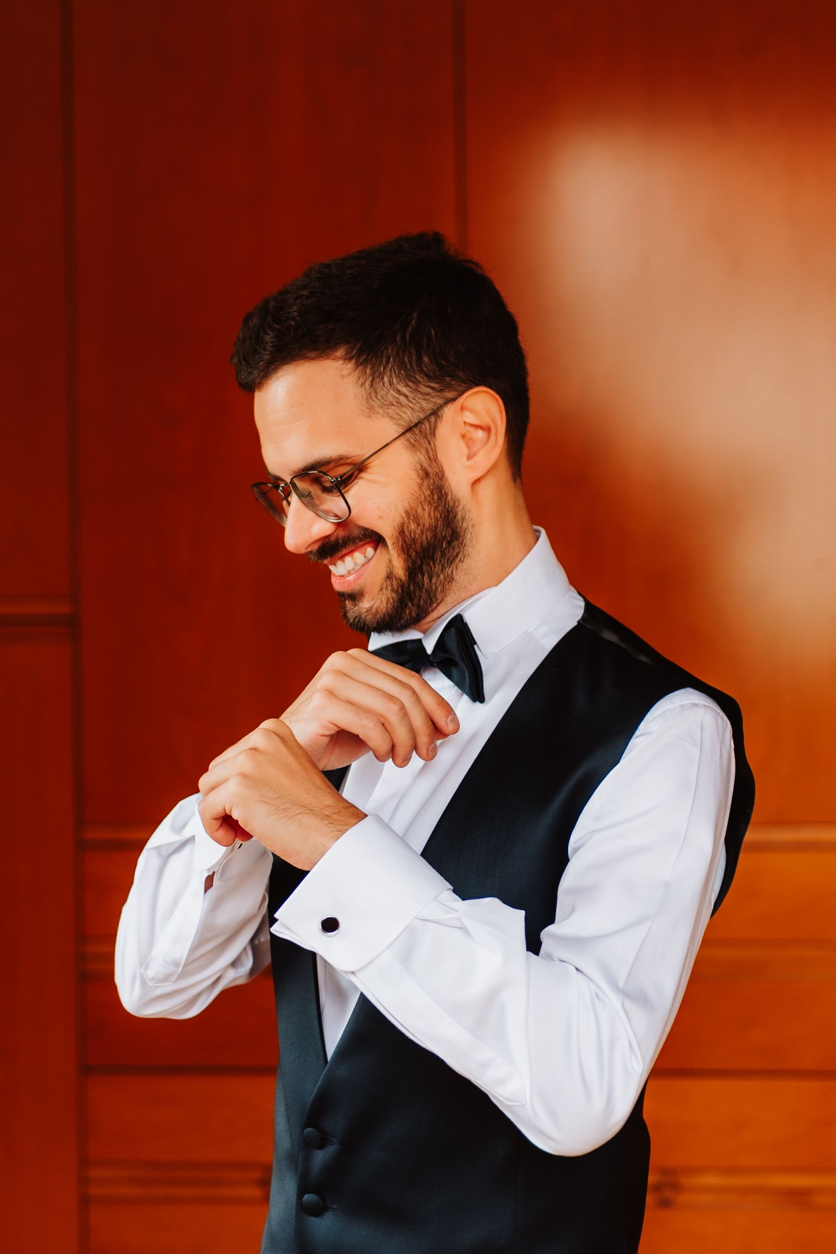 A person in a white dress shirt and black vest laughs while looking down in a warm-toned studio setting.