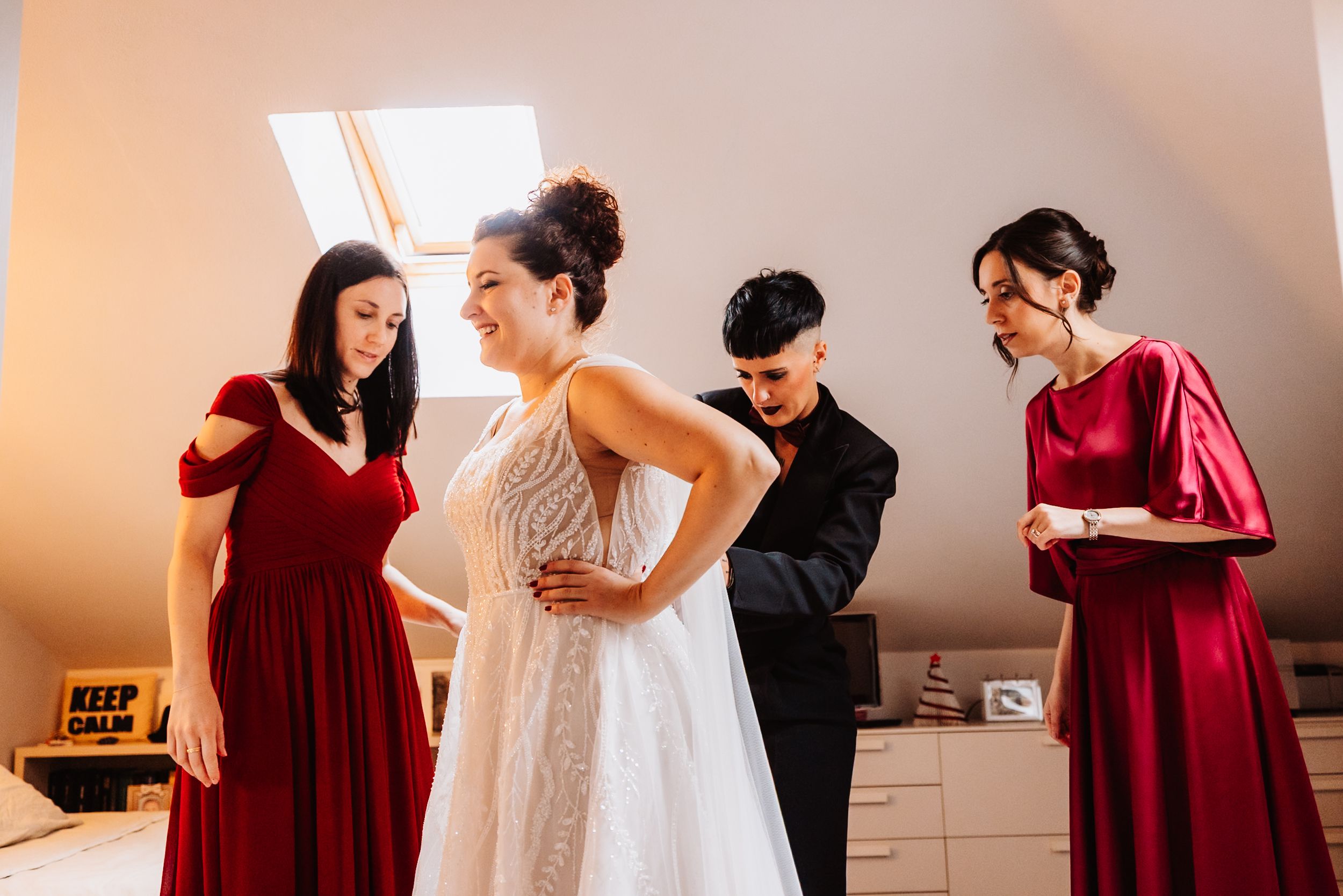 Bridesmaids in burgundy dresses help bride get ready in sunlit room before wedding ceremony.
