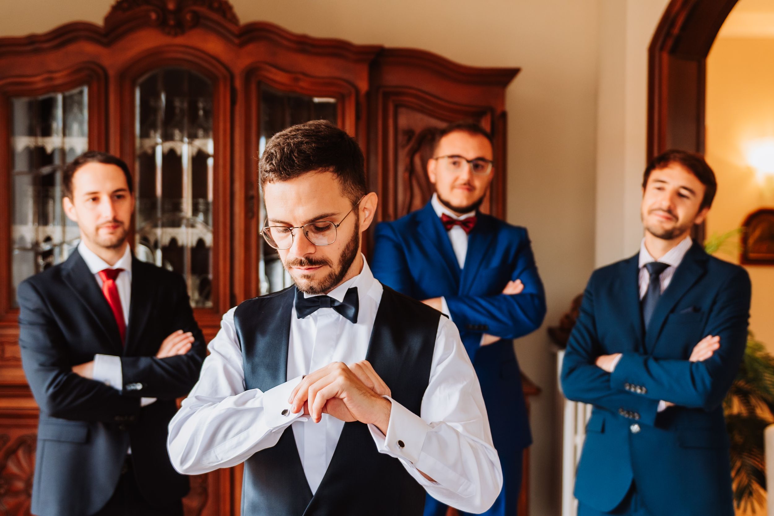 A wedding party gets ready in a formal room with groomsmen in navy suits and black bowties.