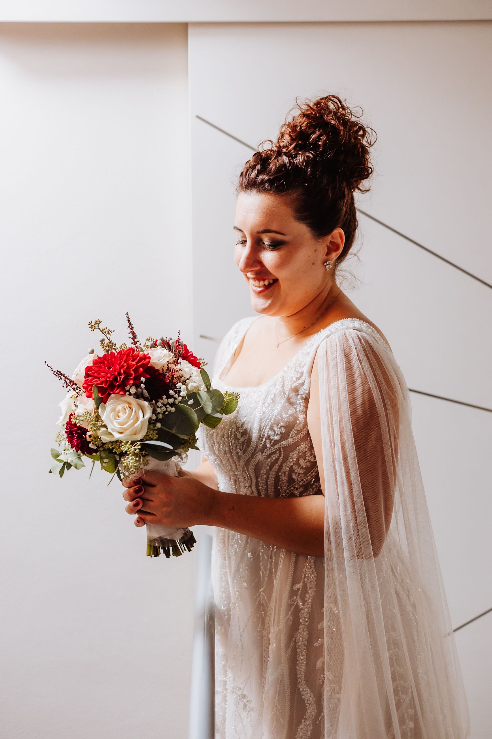 A bride in a flowing white dress with sheer sleeves holds a red and white floral bouquet against a light backdrop.