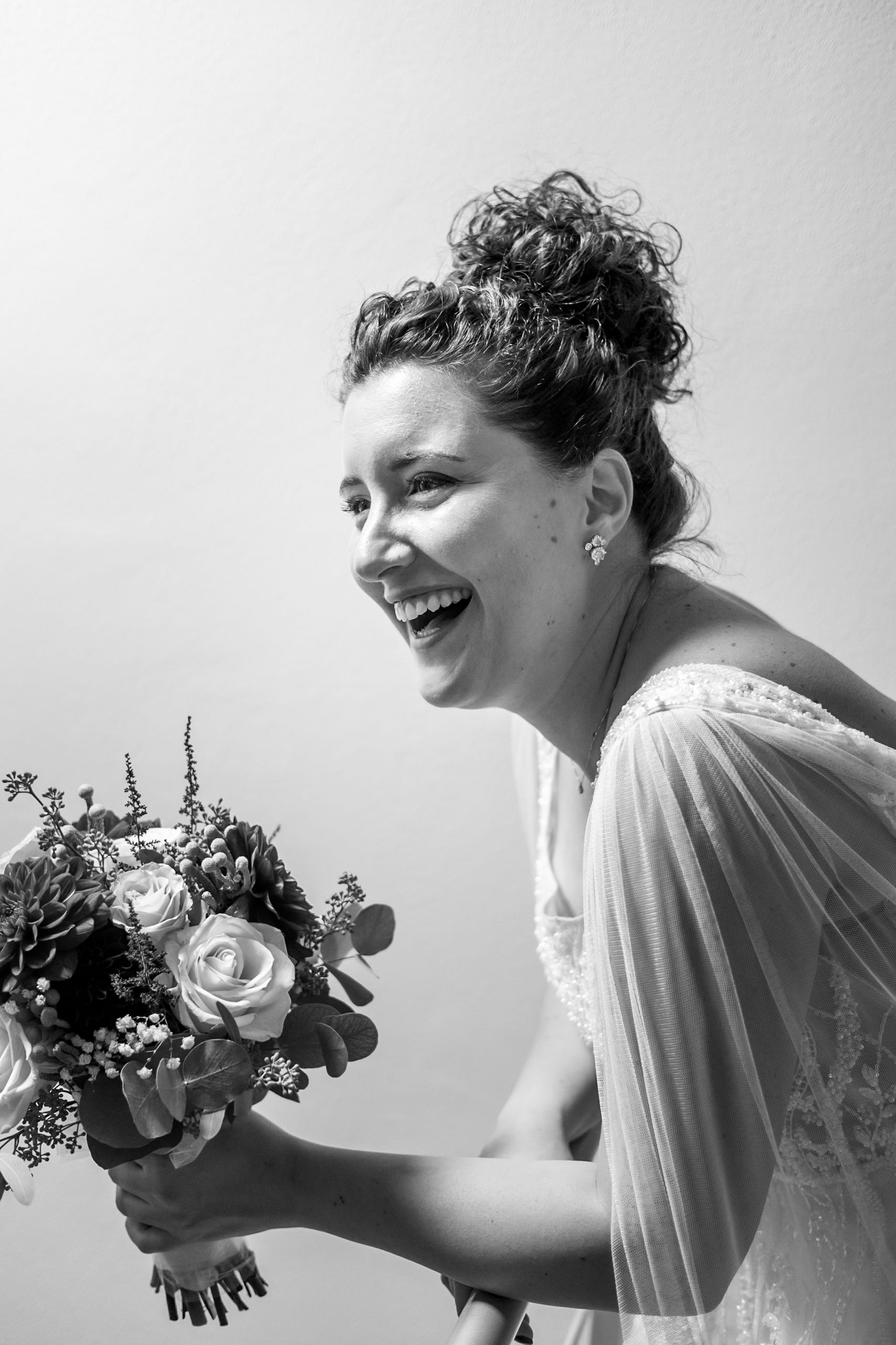 A joyful bride in a white dress laughs while holding a beautiful bouquet of roses and mixed flowers in black and white.